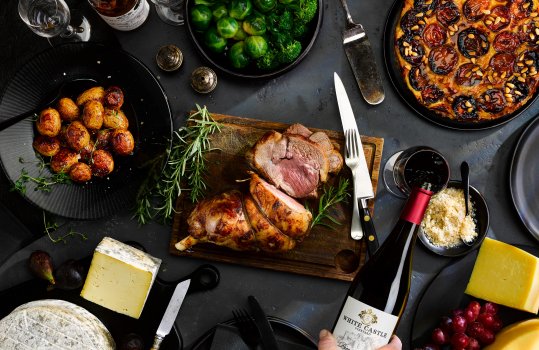 Aerial food photograph of a dinner party set made up of Welsh PGI ingredients, roast leg of Gower Salt Marsh lamb, a Denbigh plum tart, traditional Welsh Caerphilly cheeses, roast Welsh potatoes and a bottle of PGI Welsh wine being poured into a glass, all shot on dark or black tableware on a black slate background