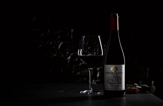 Atmospheric drinks photograph of a bottle and glass of PGI Welsh Pinot Noir wine, shot on a black slate background against a black brick wall