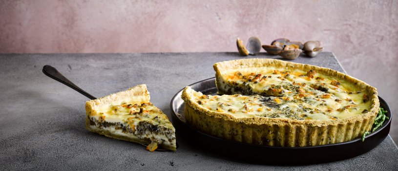 Food photograph close up of a homemade Welsh PGI laverbread and cockle quiche, with crisp homemade laverbread pastry and plump fresh cockles, shot on a black plate against a grey and pink background with cracked cockles