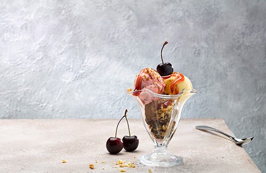 Food photograph of an ice cream sundae featuring scoops of Welsh ice creams in chocolate, strawberry and vanilla flavours, topped with strawberry sauce, chopped nuts and fresh cherries