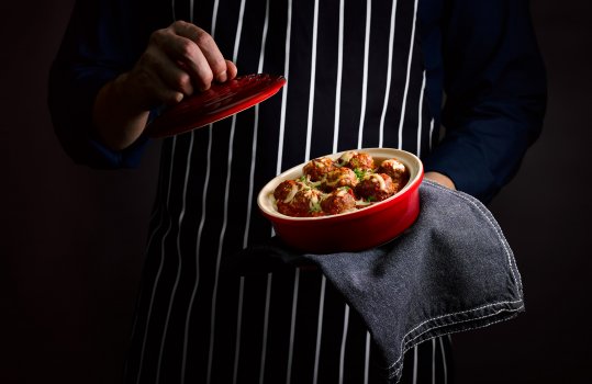 Food photograph of a man’s torso, holding a two portion dish of spicy meatballs in a rich glossy tomato sauce and topped with shredded cheese and finely chopped parsley