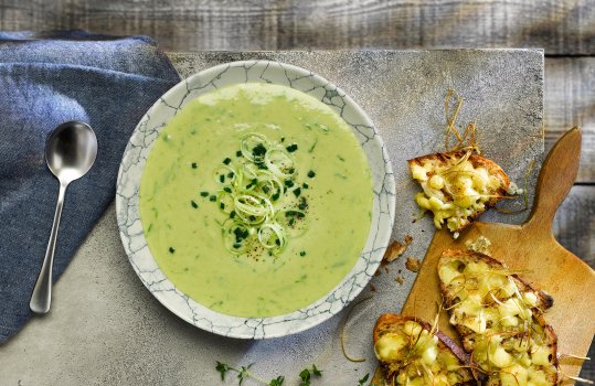 Aerial food photograph of a bowl of Welsh leek soup topped with leek rings and chopped chives. Shot on a dark grey tabletop alongside Caerphilly cheese on toast with crispy sliced leeks