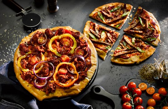 Close up food photograph of two vegan pizzas, one in the foreground topped with vegan chorizo, mixed peppers and red onion rings; and another in the background topped with mock duck strips, sesame seeds and sliced spring onions