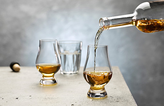 Drinks photograph close up of Welsh whisky being poured from a bottle into a traditional whisky nosing glass, a second glass with a measure of whisky is in the background. Shot on a light grey stone background