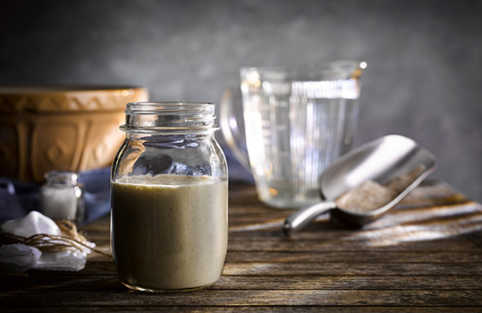 Food photograph close up of a sourdough starter in a glass jar alongside various baking tools such as a jug of water, a metal flour scoop and a large mixing bowl