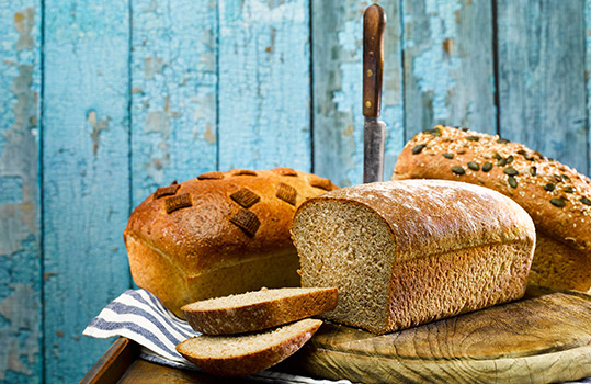 Food photograph of three homemade wholemeal loaves, one plain shown sliced, one topped with seeds and one topped with malted Shreddies cereal