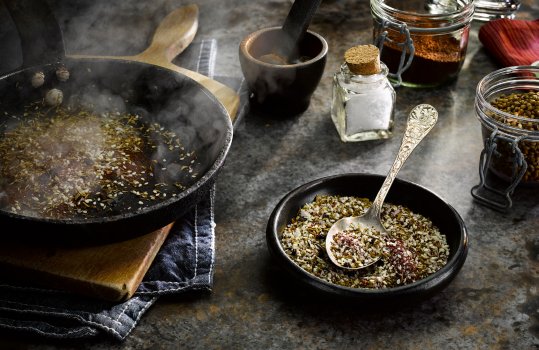 Food photograph close up of a ramekin of homemade za’atar spice blend, with a hot pan filled with spices toasting to one side, and a pestle and mortar with spice jars in the background