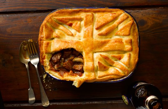 Food photograph of a beef and ale pie with a Union Flag made from pastry decorating the lid, with one corner removed to show the beef filling. Shot on a dark wooden background with vintage cutlery.