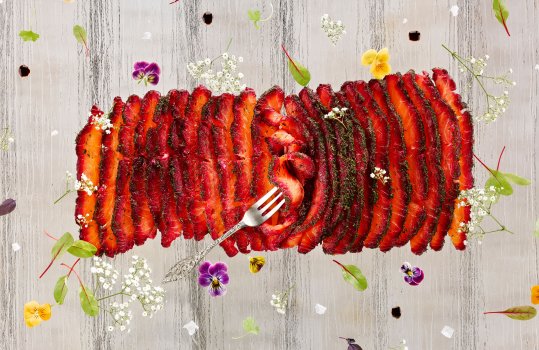 Aerial photograph of salmon gravadlax cured in beetroot and topped with dill. The whole side of cured salmon is sliced and laid out on a glass background with edible flowers and micro herbs