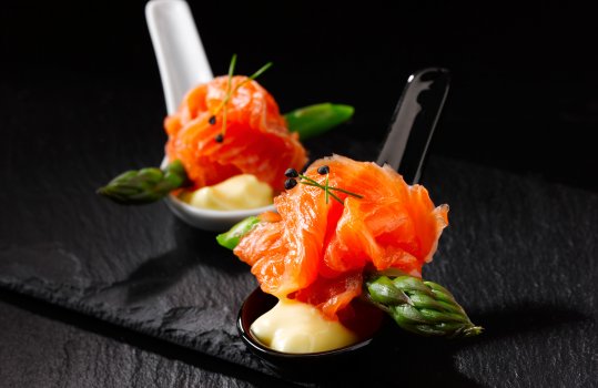 Food photograph of a pair of asian soup spoons containing a canapé of asparagus tips, cured salmon and hollandaise sauce. Shot on a black background