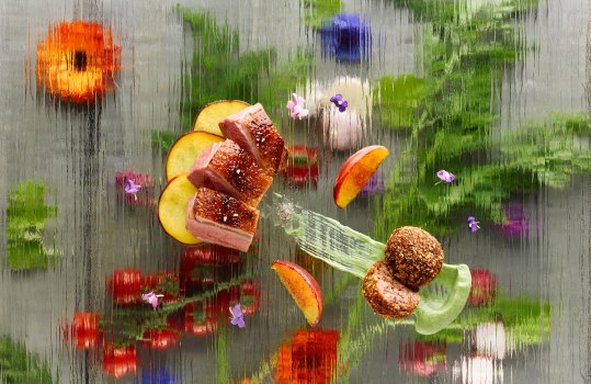 Aerial food photograph of a fine dining dish consisting of pan fried duck breast, duck leg croquette and grilled peaches. Shot on a glass background with flowers underneath