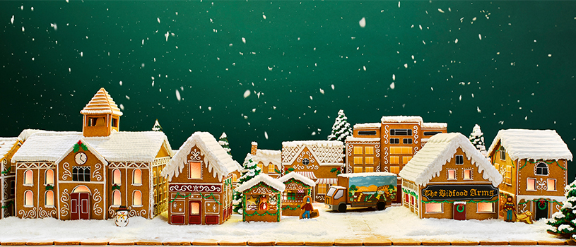 Photograph of a gingerbread village, including a pub and a schoolhouse, as well as a gingerbread delivery truck. Snow is falling and also covers the buildings and the road.