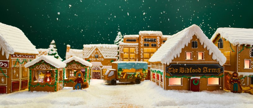 Photograph of a gingerbread village, including a pub and a schoolhouse, as well as a gingerbread delivery truck. Snow is falling and also covers the buildings and the road.