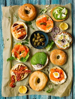 Food photograph of a breakfast bagel sharing table, featuring golden shiny toasted bagels topped with sundried tomato and grilled halloumi, smoked salmon and cream cheese, sliced avocado, crispy bacon and grilled halloumi, and egg salad. Served on baking paper garnished with parsley, sliced spring onion, wedges of lemon, and a bowl of caperberries, served on a bright blue wooden tabletop