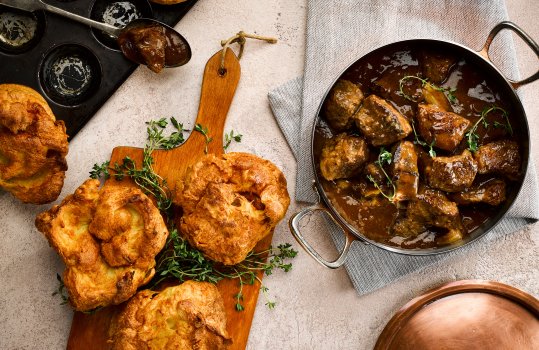 Food photograph of beef stew with Yorkshire puddings, a saucepan full of rich shiny beef stew, alongside crisp fluffy homemade Yorkshire puddings served on a wooden board with sprigs of thyme to garnish, all served on a grey tabletop with a blue and white striped tea towel