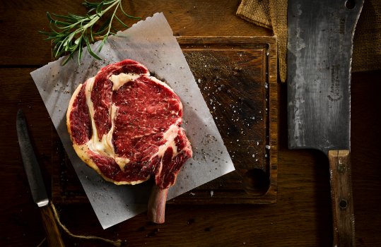 Aerial food photograph of a shiny raw bone in rib eye beef steak, served on butchers paper on top of a dark wooden board, garnished with sea salt and cracked black pepper, with a sprig of rosemary, served on a dark wooden table with a small knife, a large cleaver, butchers string and a hessian cloth