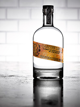 Drinks photograph of four bottles of small batch gin, each shown individually on a reflective metal tabletop with white subway tiles in the background