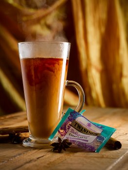 Food photograph of a tall glass cup of chai spiced tea, served on a wooden tabletop with whole cinnamon and star anise, with a gold curtain in the background. The cup of chai is steaming and milk is swirling into the tea