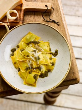 Food photograph of a bowl of ravioli topped with parmesan shavings and pesto, served on a wooden farmhouse table with a wedge of parmesan; with the table corner and a leg visible, on top of white floorboards.