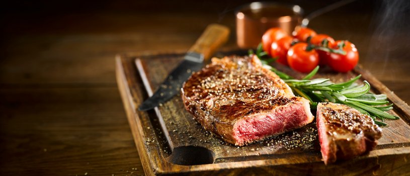 Food photograph of a juicy rare beef sirloin steak, with a crunchy crisp experior and a juicy pink inside. Served on a wooden board with rosemary and vine cherry tomatoes, on a wooden tabletop