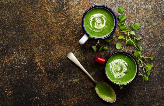 Aerial food photograph of watercress soup served in enamel cups and finished with a swirl of cream and cracked pepper, served alongside watercress for garnish, on a paint speckled dark tabletop