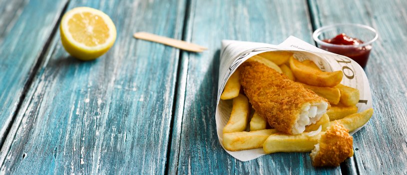 Aerial food photograph of takeaway fish and chips, crisp golden battered cod fillet served in newspaper on top of golden chips, alongside a pot of tomato ketchup, wedges of lemon, and a wooden chip fork. Served on a blue wooden tabletop