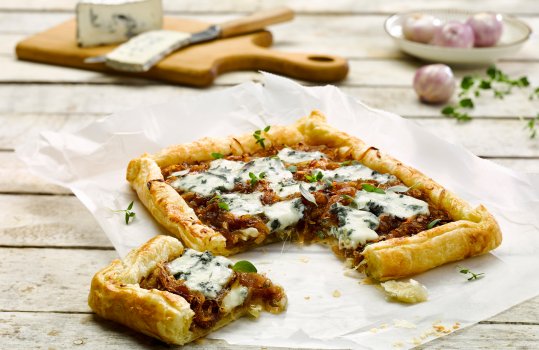 Food photograph of a Dol Las (soft blue sheep cheese) and caramelised onion puff pastry tart. One slice has been cut from the tart to show the filling in more detail