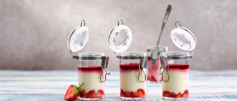 Food photograph of mini dessert pots, white chocolate mousse with strawberries, topped with strawberry syrup served in miniature jars on a painted wooden tabletop, garnished with half a strawberry